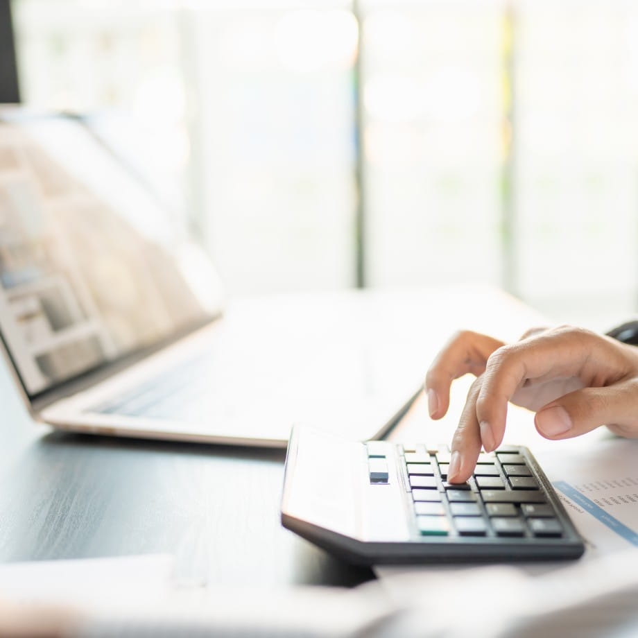 Close-up of a person using a calculator next to a laptop at a bright office desk
