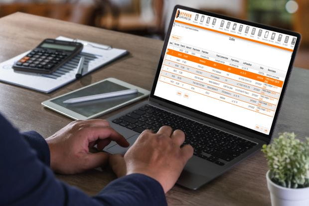 Contractor working on a laptop showing a SupplyPro GM order schedule, with a tablet and calculator on the desk